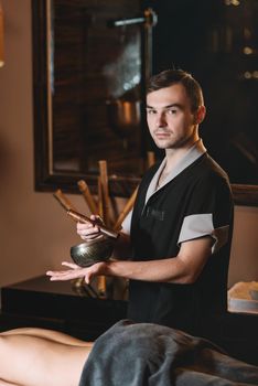 Specialist holding in hands Brass Hammer Singing Bowl. Young charming girl on a panchakarma procedure laying on a massage table. beautiful woman spending time at modern spa cabinet relaxing. Soft yellow light.