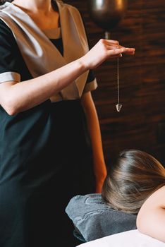 Specialist making biolocation with crystal pendulum. Young charming girl on a panchakarma procedure laying on a massage table. beautiful woman spending time at modern spa cabinet relaxing. Soft yellow light.