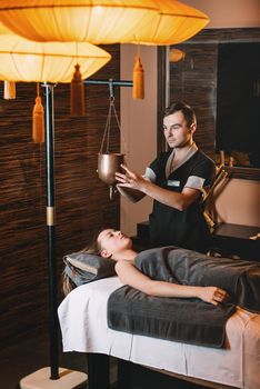 Specialist making shirodhara massage form of ayurveda therapy. Young charming girl on a panchakarma procedure laying on a massage table. beautiful woman spending time at modern spa cabinet relaxing. Soft yellow light.