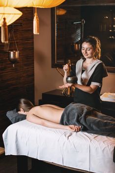 Specialist holding in hands Brass Hammer Singing Bowl. Young charming girl on a panchakarma procedure laying on a massage table. beautiful woman spending time at modern spa cabinet relaxing. Soft yellow light.