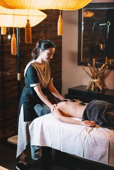 Specialist making hot stone massage to a client. Young charming girl on a panchakarma procedure laying on a massage table. beautiful woman spending time at modern spa cabinet relaxing. Soft yellow light.