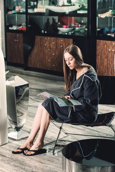 Portrait of young beautiful woman relaxing in a chair waiting for a procedure. in a bathrobe with magazine in hands. Luxery spa center.