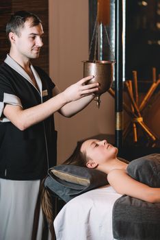 Specialist making shirodhara massage form of ayurveda therapy. Young charming girl on a panchakarma procedure laying on a massage table. beautiful woman spending time at modern spa cabinet relaxing. Soft yellow light.