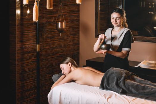 Specialist holding in hands Brass Hammer Singing Bowl. Young charming girl on a panchakarma procedure laying on a massage table. beautiful woman spending time at modern spa cabinet relaxing. Soft yellow light.