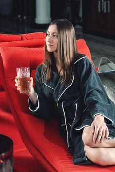 Portrait of young beautiful woman relaxing in a fashionable red chair in a bathrobe with a detox drink in a hand. Luxery spa center