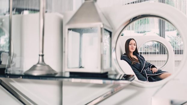 Portrait of young beautiful woman relaxing in a fashionable white sofa in a gray bathrobe, waiting for treatment. Luxery spa center