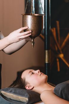 Specialist making shirodhara massage form of ayurveda therapy. Young charming girl on a panchakarma procedure laying on a massage table. beautiful woman spending time at modern spa cabinet relaxing. Soft yellow light.