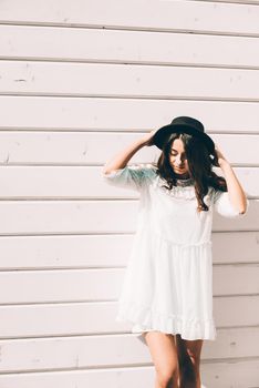 Sunny lifestyle fashion portrait of young stylish hipster woman walking on the street, wearing trendy white dress, black hat and boots. White wooden backgrond.