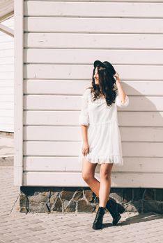 Sunny lifestyle fashion portrait of young stylish hipster woman walking on the street, wearing trendy white dress, black hat and boots. White wooden backgrond.