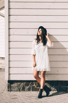 Sunny lifestyle fashion portrait of young stylish hipster woman walking on the street, wearing trendy white dress, black hat and boots. White wooden backgrond.