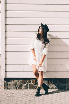 Sunny lifestyle fashion portrait of young stylish hipster woman walking on the street, wearing trendy white dress, black hat and boots. White wooden backgrond.