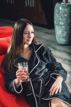 Portrait of young beautiful woman relaxing in a fashionable red chair in a bathrobe with a detox drink in a hand. Luxery spa center