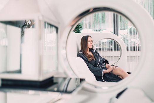 Portrait of young beautiful woman relaxing in a fashionable white sofa in a gray bathrobe, waiting for treatment. Luxery spa center