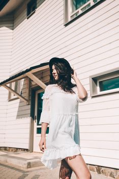 Sunny lifestyle fashion portrait of young stylish hipster woman walking on the street, wearing trendy white dress, black hat and boots. White wooden house on a backgrond.