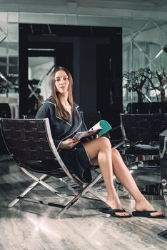 Portrait of young beautiful woman relaxing in a chair waiting for a procedure. in a bathrobe with magazine in hands. Luxery spa center.