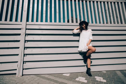 Sunny lifestyle fashion portrait of young stylish hipster woman walking on the street, wearing trendy white dress, black hat and boots. Gray wooden backgrond.