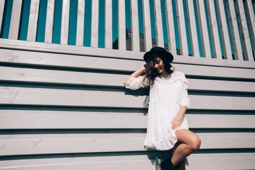 Sunny lifestyle fashion portrait of young stylish hipster woman walking on the street, wearing trendy white dress, black hat and boots. Gray wooden backgrond.