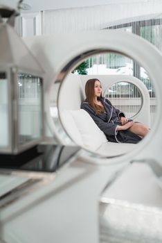 Portrait of young beautiful woman relaxing in a fashionable white sofa in a gray bathrobe, waiting for treatment. Luxery spa center
