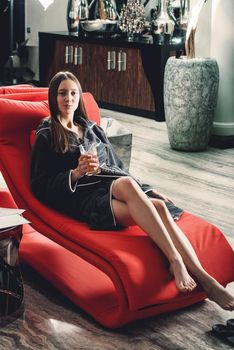 Portrait of young beautiful woman relaxing in a fashionable red chair in a bathrobe with a detox drink in a hand. Luxery spa center