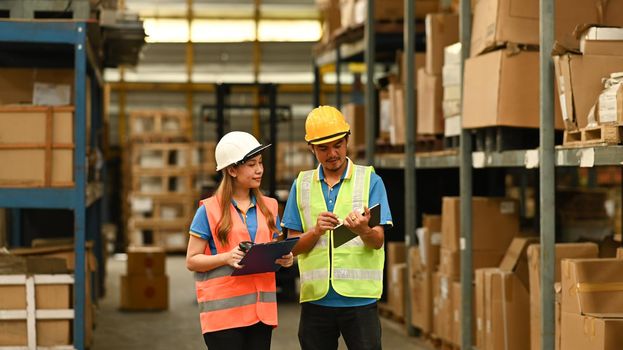 Warehouse workers wearing safety hardhat discussing work while walking in aisle between rows of tall shelves full of packed boxes.