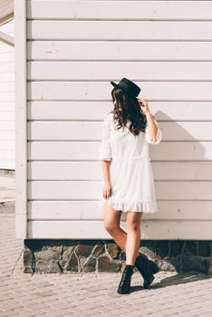 Sunny lifestyle fashion portrait of young stylish hipster woman walking on the street, wearing trendy white dress, black hat and boots. White wooden backgrond.