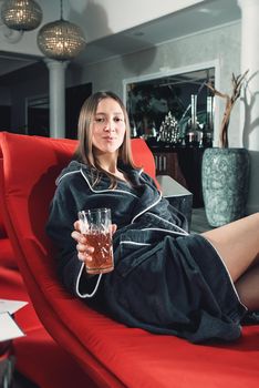 Portrait of young beautiful woman relaxing in a fashionable red chair in a bathrobe with a detox drink in a hand. Luxery spa center