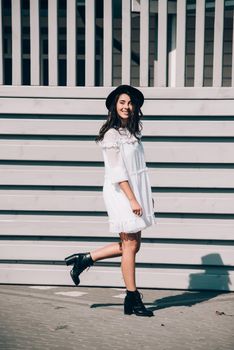 Sunny lifestyle fashion portrait of young stylish hipster woman walking on the street, wearing trendy white dress, black hat and boots. Gray wooden backgrond.