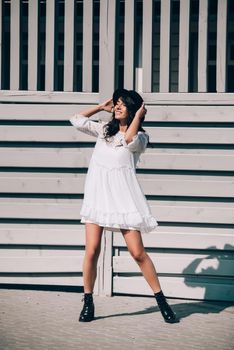 Sunny lifestyle fashion portrait of young stylish hipster woman walking on the street, wearing trendy white dress, black hat and boots. Gray wooden backgrond.