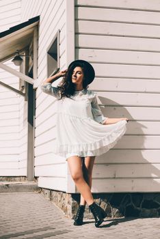 Sunny lifestyle fashion portrait of young stylish hipster woman walking on the street, wearing trendy white dress, black hat and boots. White wooden house on a backgrond.