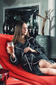 Portrait of young beautiful woman relaxing in a fashionable red chair in a bathrobe with a detox drink in a hand. Luxery spa center