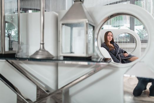 Portrait of young beautiful woman relaxing in a fashionable white sofa in a gray bathrobe, waiting for treatment. Luxery spa center