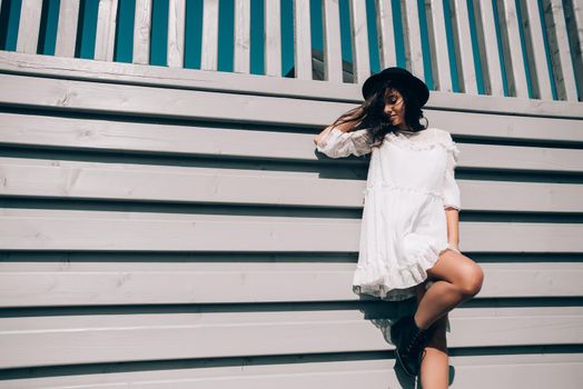 Sunny lifestyle fashion portrait of young stylish hipster woman walking on the street, wearing trendy white dress, black hat and boots. Gray wooden backgrond.