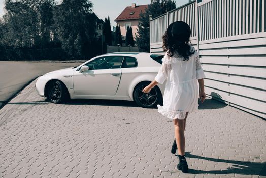 Sunny lifestyle fashion portrait of young stylish hipster woman walking to her car, wearing trendy white dress, black hat and boots. Gray wooden backgrond.