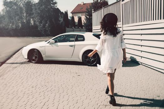 Sunny lifestyle fashion portrait of young stylish hipster woman walking to her car, wearing trendy white dress, black hat and boots. Gray wooden backgrond.