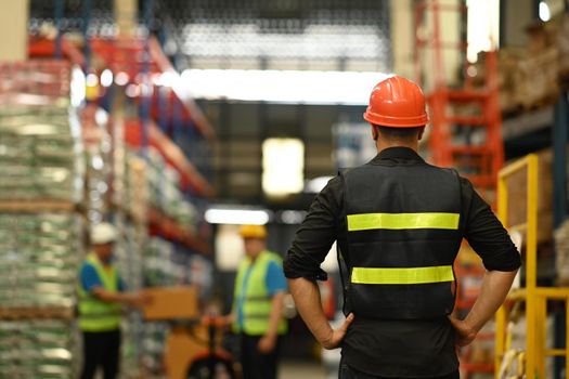 Back view of male manager wearing hardhat and reflective jacket checking inventory production stock control in warehouse.