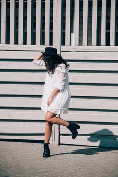 Sunny lifestyle fashion portrait of young stylish hipster woman walking on the street, wearing trendy white dress, black hat and boots. Gray wooden backgrond.