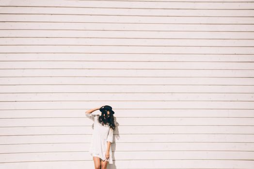 Sunny lifestyle fashion portrait of young stylish hipster woman walking on the street, wearing trendy white dress, black hat and boots. White wooden backgrond.