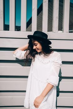 Sunny lifestyle fashion portrait of young stylish hipster woman walking on the street, wearing trendy white dress, black hat and boots. Gray wooden backgrond.