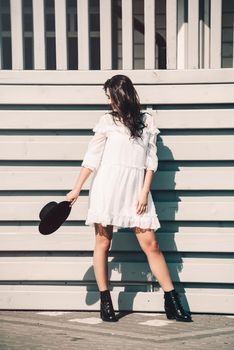 Sunny lifestyle fashion portrait of young stylish hipster woman walking on the street, wearing trendy white dress, black hat and boots. Gray wooden backgrond.