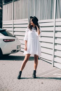 Sunny lifestyle fashion portrait of young stylish hipster woman walking on the street, wearing trendy white dress, black hat and boots. Gray wooden backgrond.