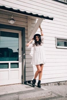 Sunny lifestyle fashion portrait of young stylish hipster woman walking on the street, wearing trendy white dress, black hat and boots. White wooden house on a backgrond.