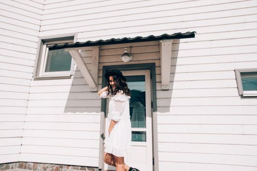 Sunny lifestyle fashion portrait of young stylish hipster woman walking on the street, wearing trendy white dress, black hat and boots. White wooden house on a backgrond.