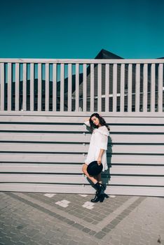 Sunny lifestyle fashion portrait of young stylish hipster woman walking on the street, wearing trendy white dress, black hat and boots. Gray wooden backgrond.