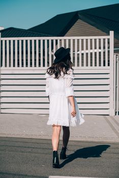 Sunny lifestyle fashion portrait of young stylish hipster woman walking on the street, wearing trendy white dress, black hat and boots. Gray wooden backgrond.