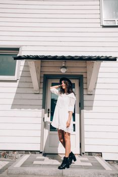 Sunny lifestyle fashion portrait of young stylish hipster woman walking on the street, wearing trendy white dress, black hat and boots. White wooden house on a backgrond.