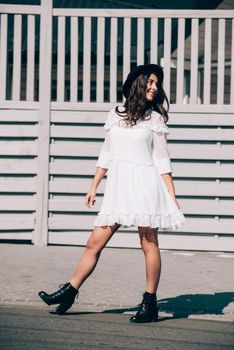 Sunny lifestyle fashion portrait of young stylish hipster woman walking on the street, wearing trendy white dress, black hat and boots. Gray wooden backgrond.