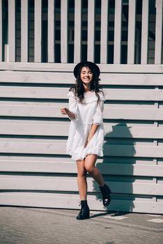 Sunny lifestyle fashion portrait of young stylish hipster woman walking on the street, wearing trendy white dress, black hat and boots. Gray wooden backgrond.