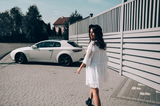 Sunny lifestyle fashion portrait of young stylish hipster woman walking to her car, wearing trendy white dress, black hat and boots. Gray wooden backgrond.