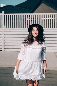 Sunny lifestyle fashion portrait of young stylish hipster woman walking on the street, wearing trendy white dress, black hat and boots. Gray wooden backgrond.