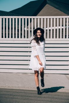 Sunny lifestyle fashion portrait of young stylish hipster woman walking on the street, wearing trendy white dress, black hat and boots. Gray wooden backgrond.